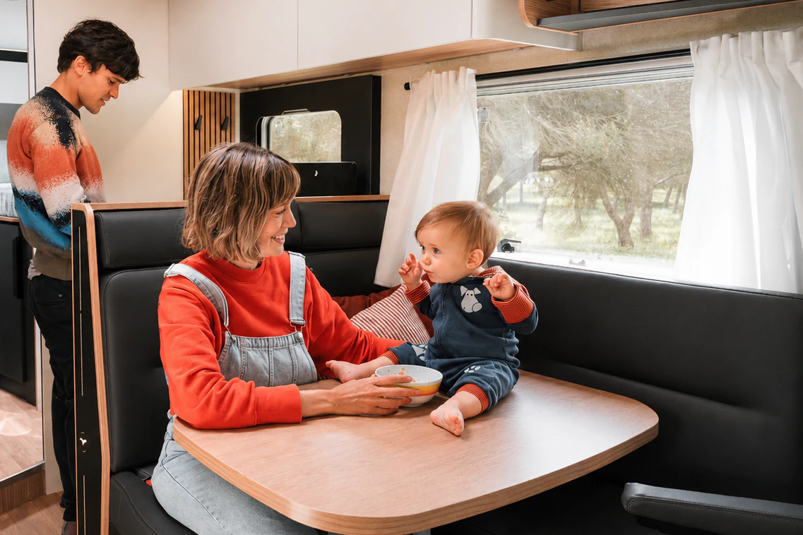 Van interior with dining table, black bench seats, overhead cabinets, large window with curtains, and a side kitchen area.
