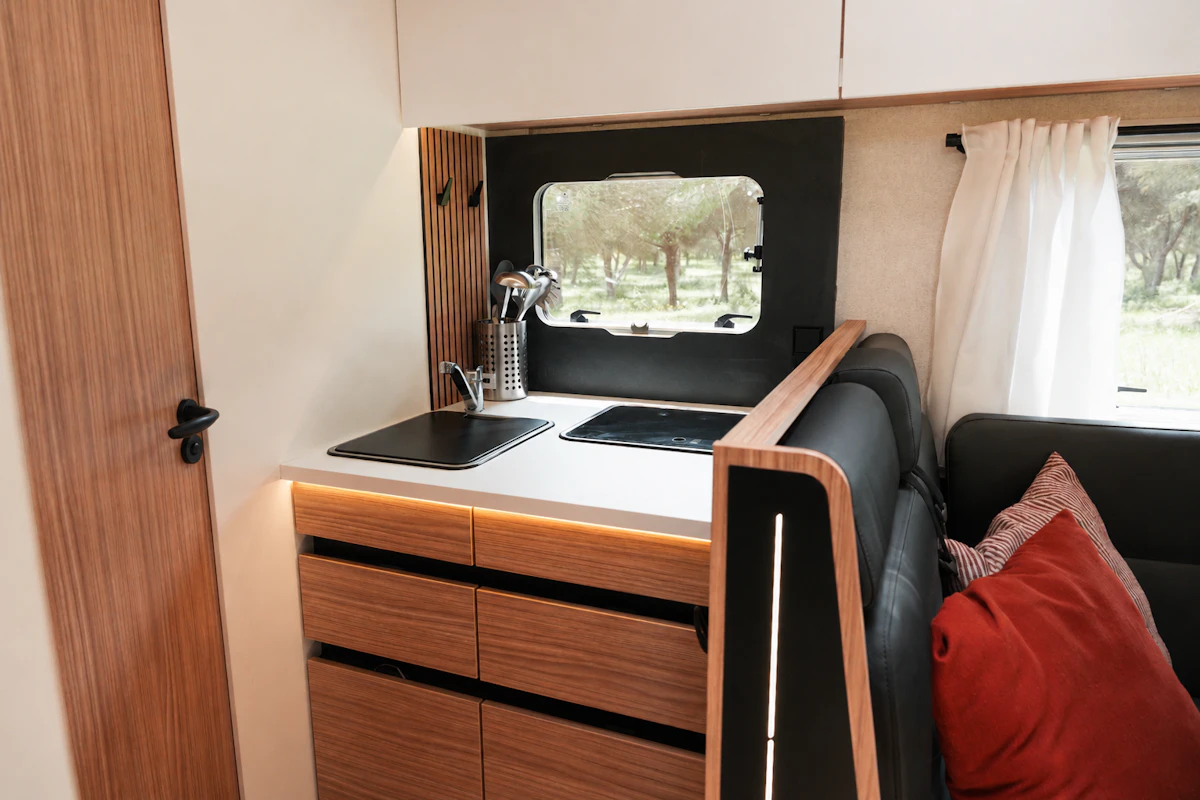 Compact van kitchen with sink, stove, wooden cabinets, black sofa, red cushion, and window above countertop.
