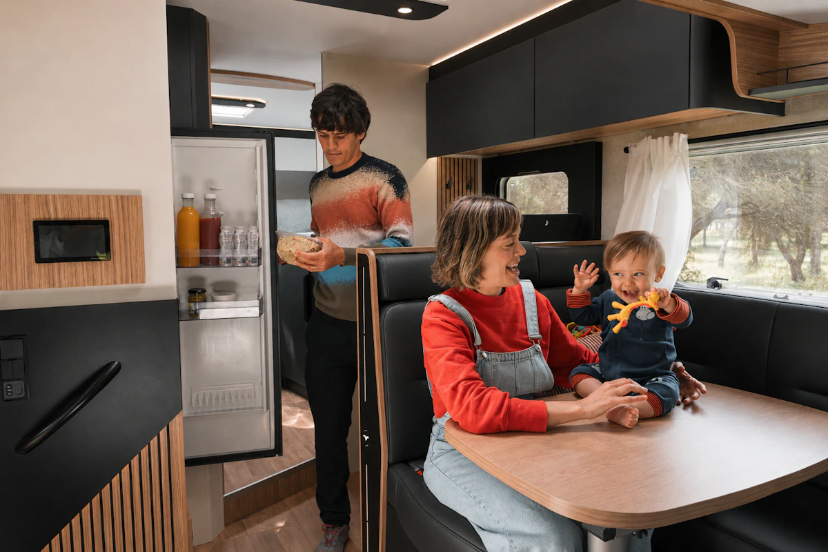 Van interior with dining table, bench seats, overhead cabinets, open fridge, and visible kitchen counter area.
