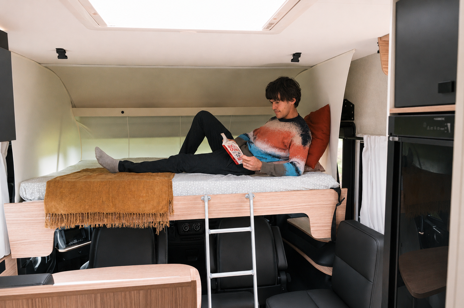 Van interior with elevated bed, ladder, seating area below, blanket, pillow, and visible storage and kitchen units.