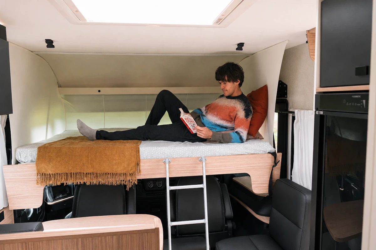 Van interior with elevated bed, ladder, seating area below, blanket, pillow, and visible storage and kitchen units.