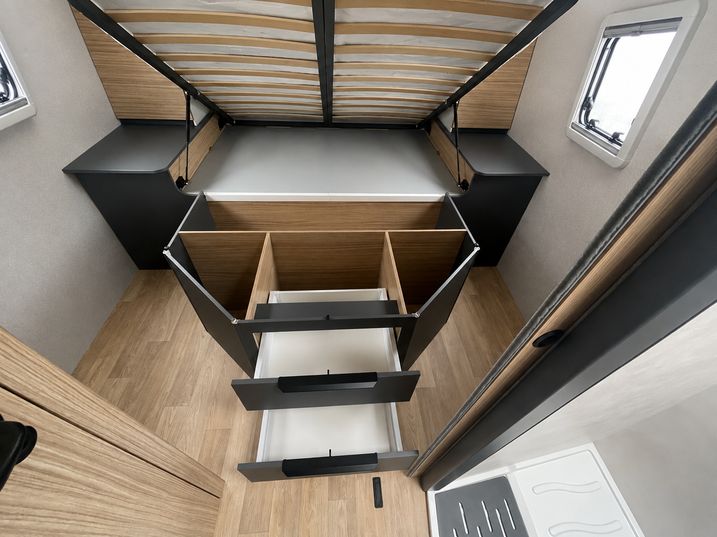 Van interior with wood flooring, open storage drawers, bed frame lifted, side cabinets, and two windows visible.