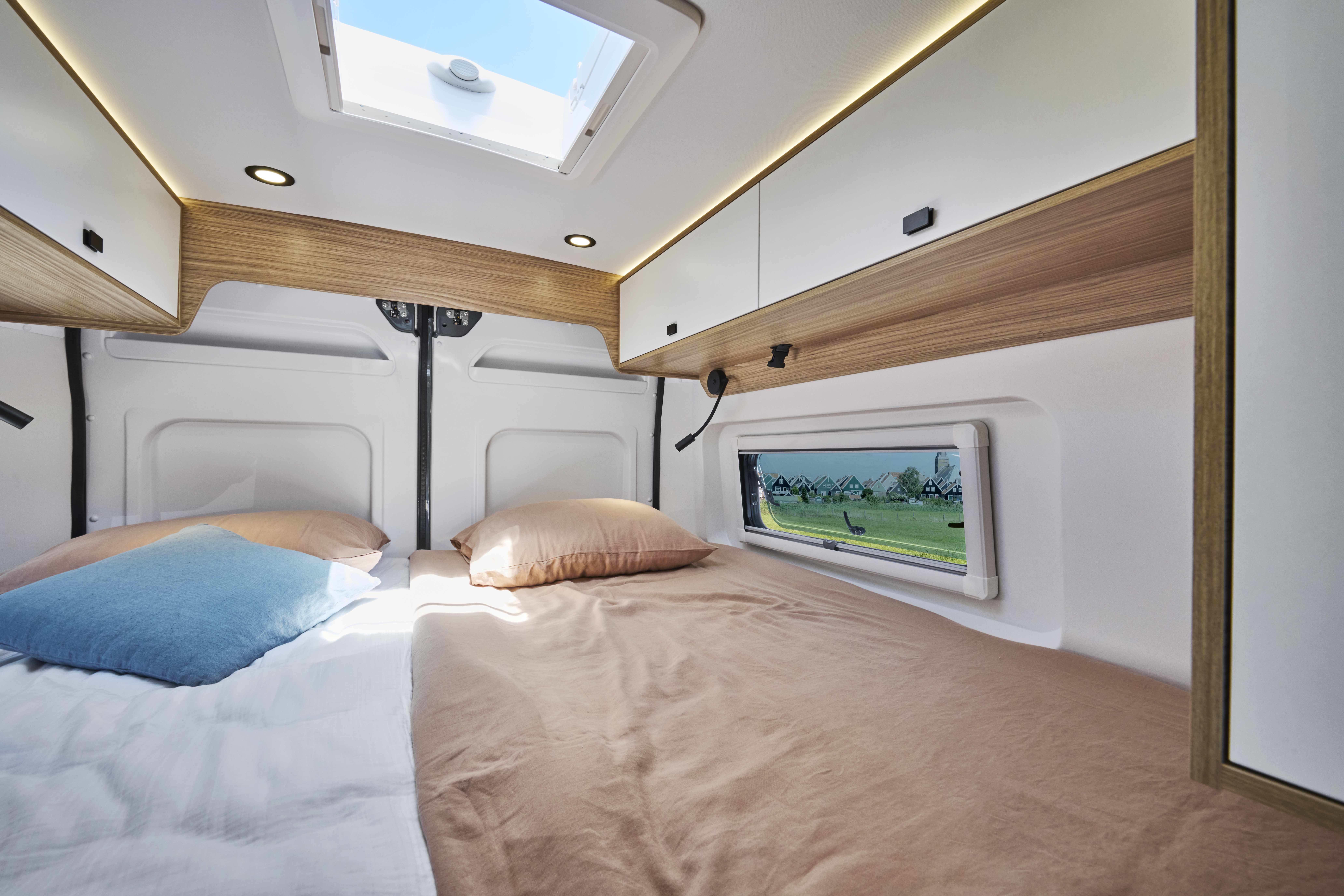 Van interior with double bed, overhead cabinets, side window, ceiling light, and skylight visible.