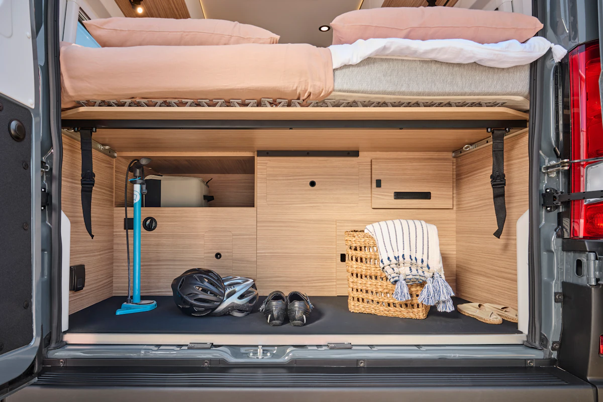 Rear interior view of van showing storage space, cabinets, bedding above, and various equipment.