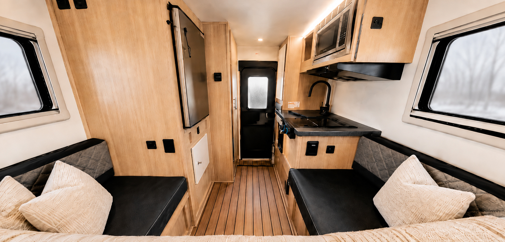 Van interior with two benches, kitchen area with sink and microwave, wood paneling, and rear door visible.