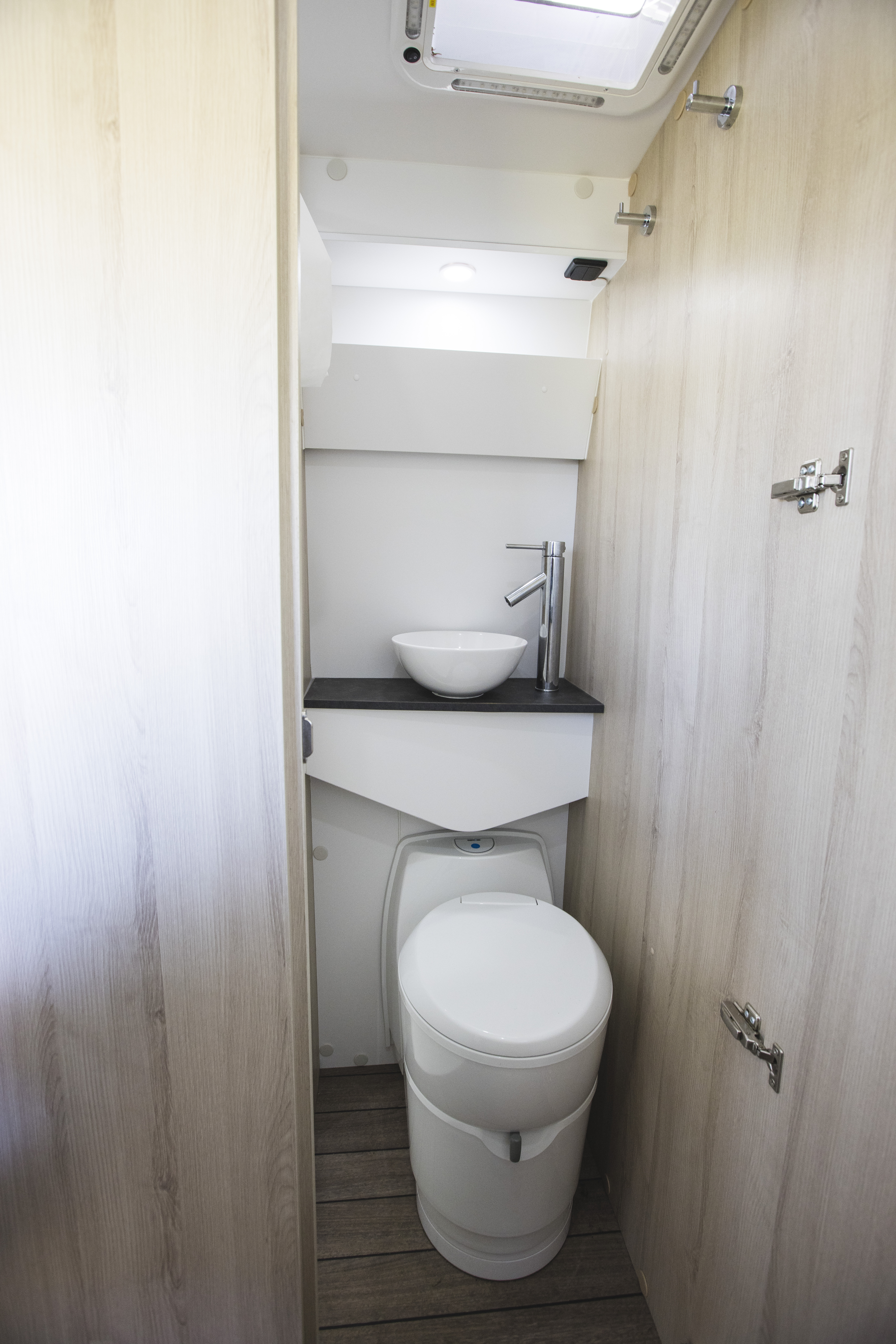Compact camper van bathroom with a white toilet, wooden floor, and light wood panels on both sides.