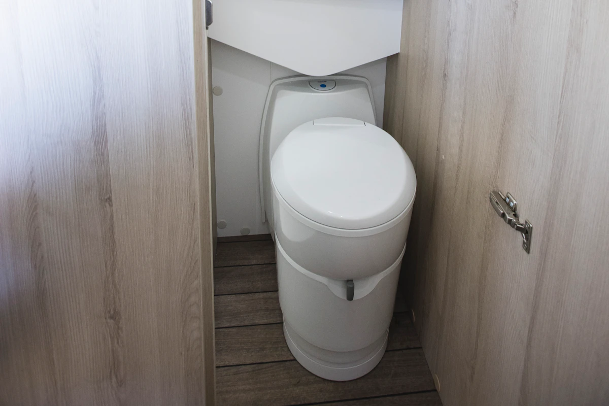 Compact camper van bathroom with a white toilet, wooden floor, and light wood panels on both sides.