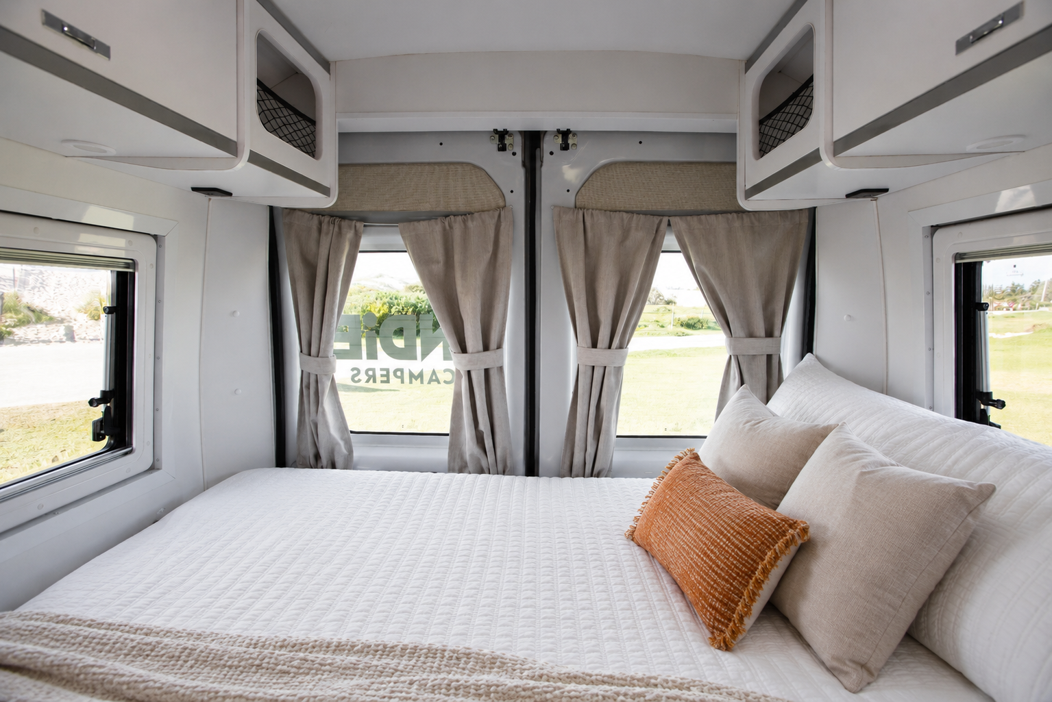 Van interior with rear bed, pillows, overhead storage cabinets, and windows with curtains on three sides.