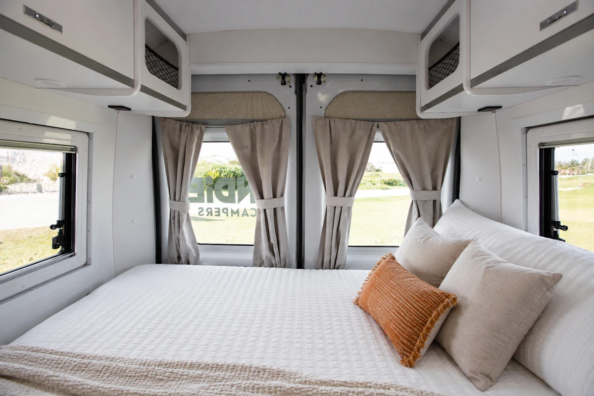 Van interior with rear bed, pillows, overhead storage cabinets, and windows with curtains on three sides.