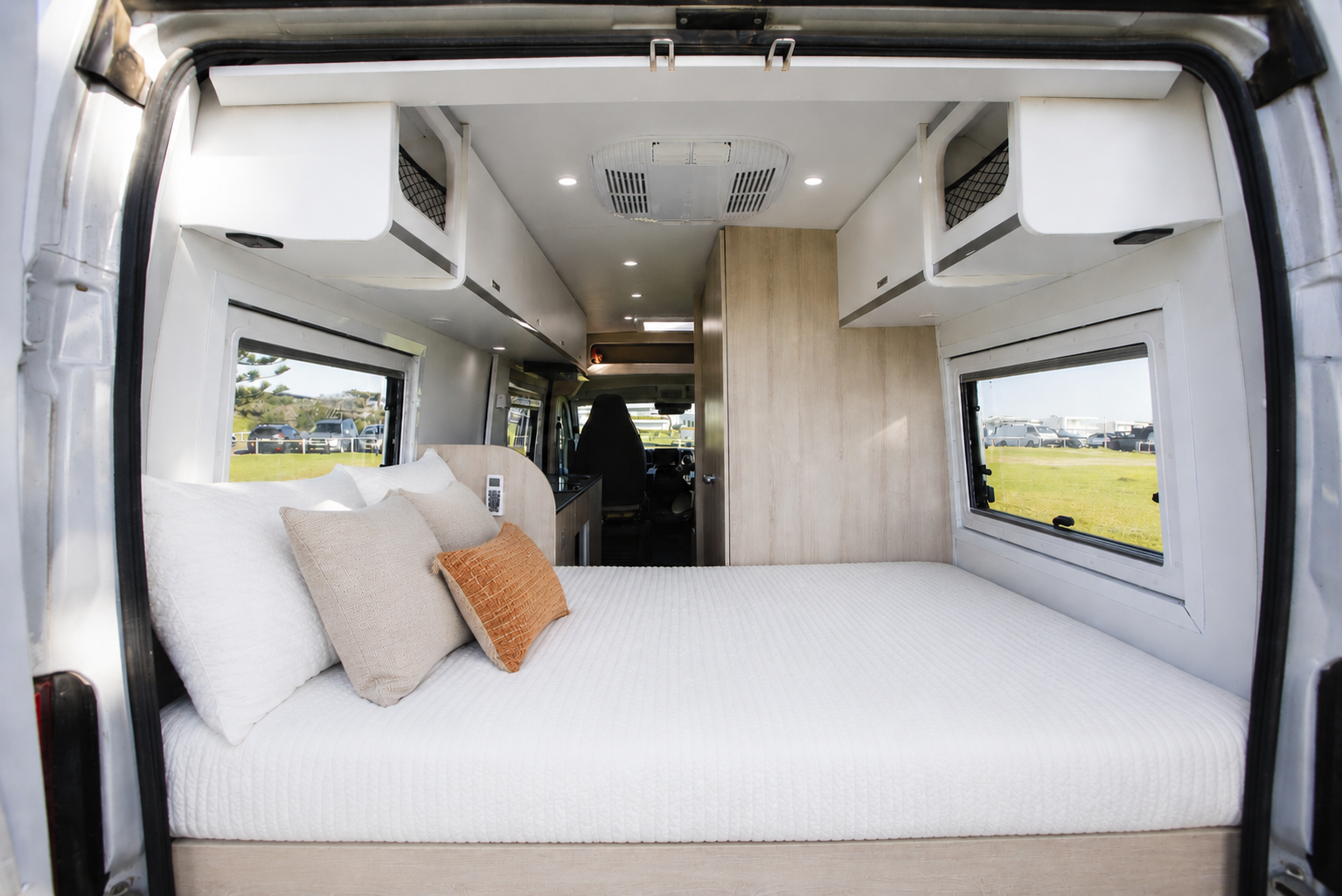 Rear view of van interior showing bed, overhead storage, windows, pillows, and partial kitchen area.