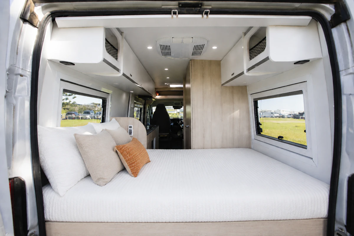 Rear view of van interior showing bed, overhead storage, windows, pillows, and partial kitchen area.