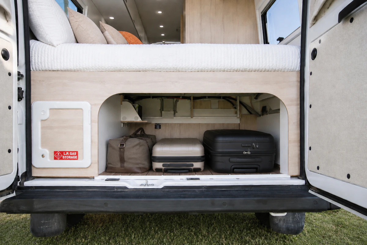 Rear view of van interior showing storage area with bags and bed above, shelves and light wood cabinets visible.