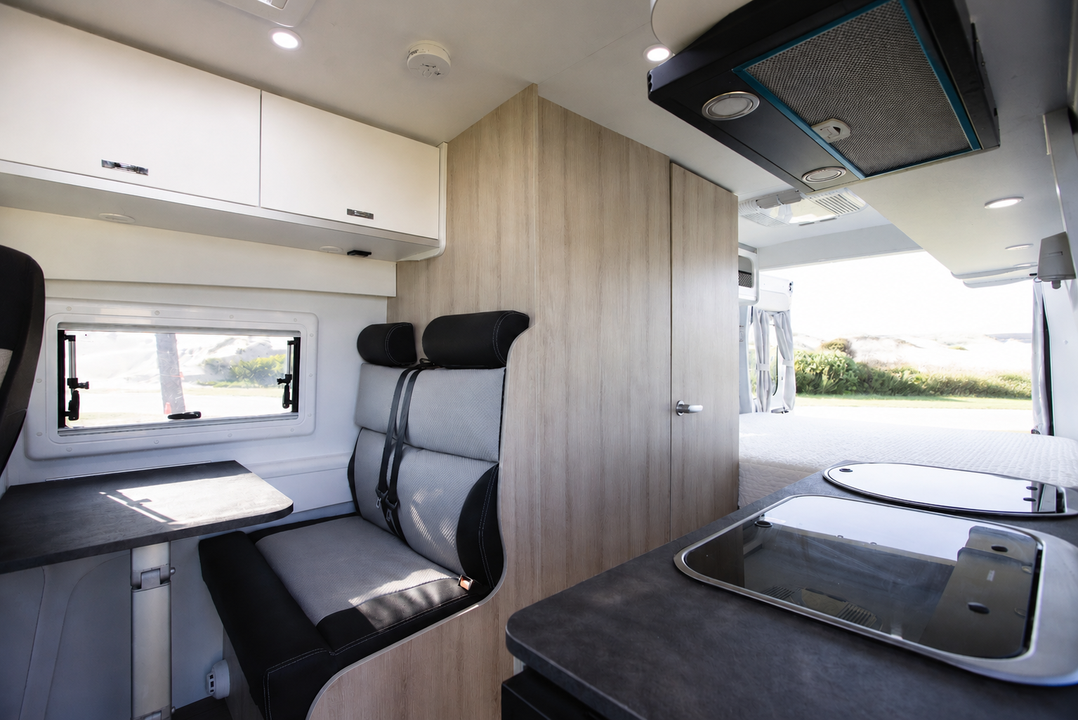 Van interior with seating area, table, overhead storage, kitchenette with sink and stove, and wooden partition wall.