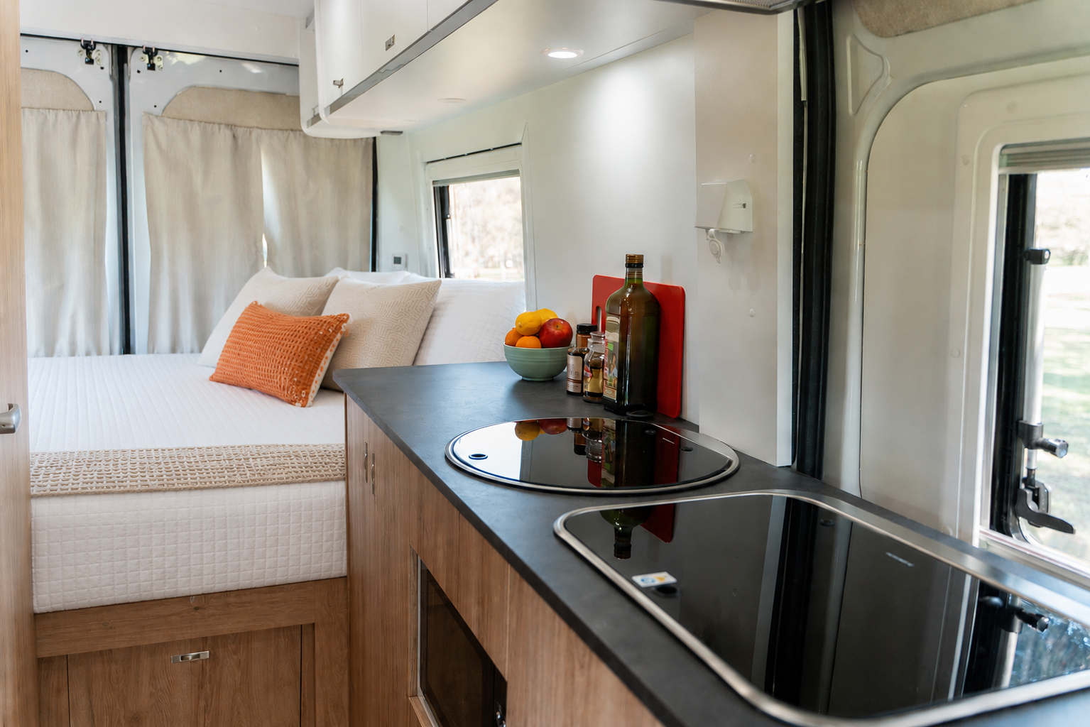 Van interior with bed, kitchenette, induction stove, sink, overhead cabinets, and assorted kitchen essentials on the counter.