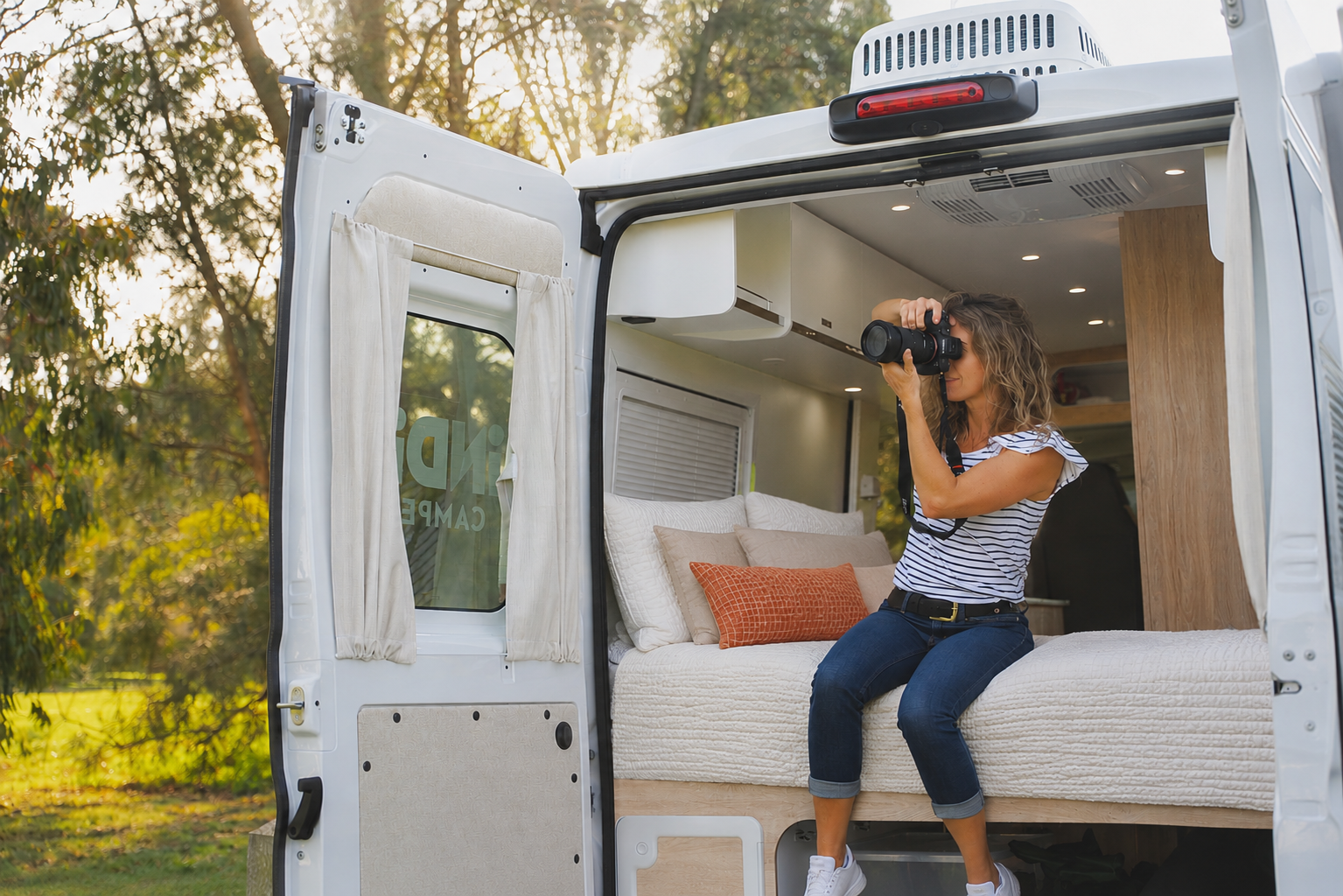 Van interior with open rear doors, visible bed, pillows, overhead storage, ceiling lights, and wooden partition.