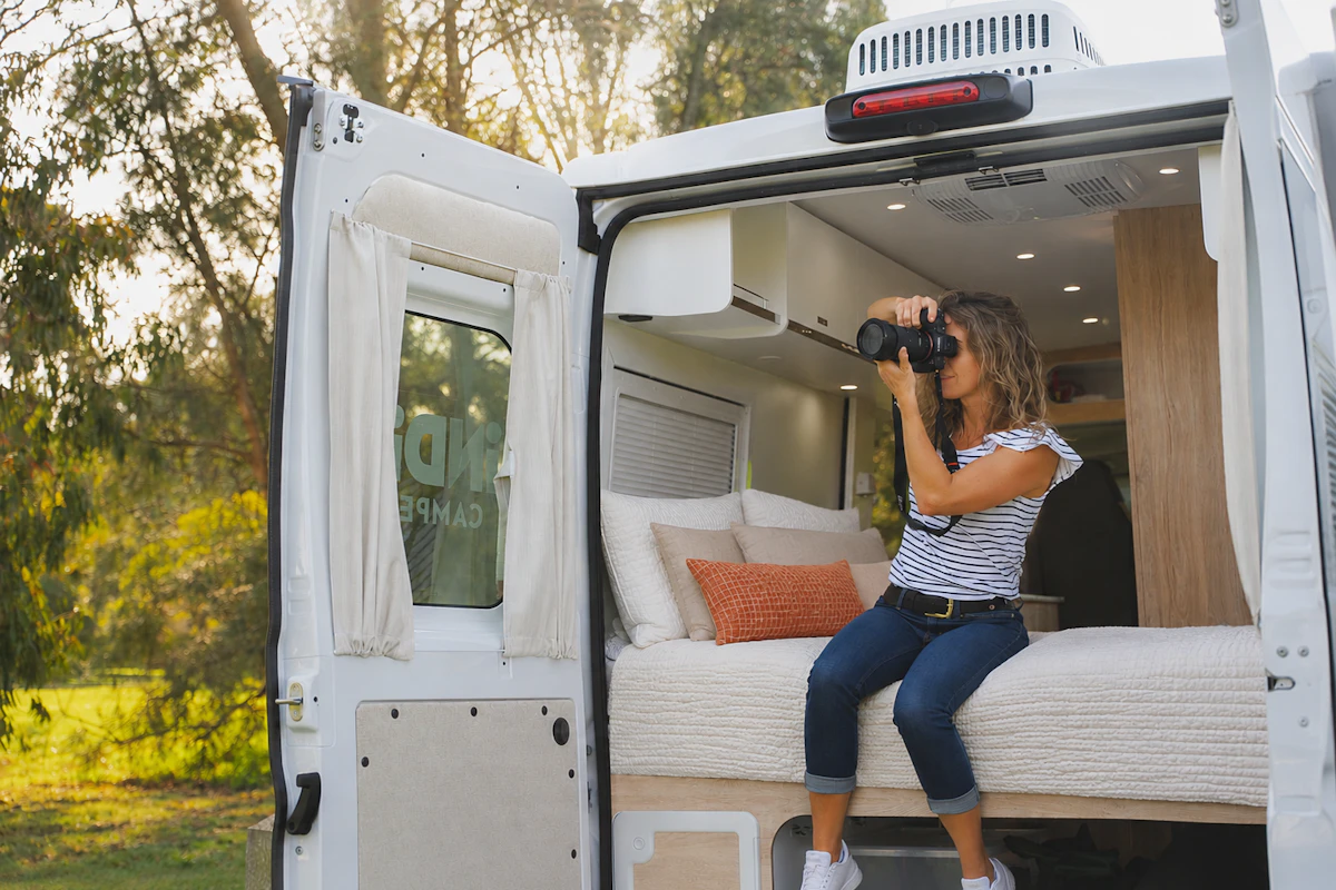 Van interior with open rear doors, visible bed, pillows, overhead storage, ceiling lights, and wooden partition.