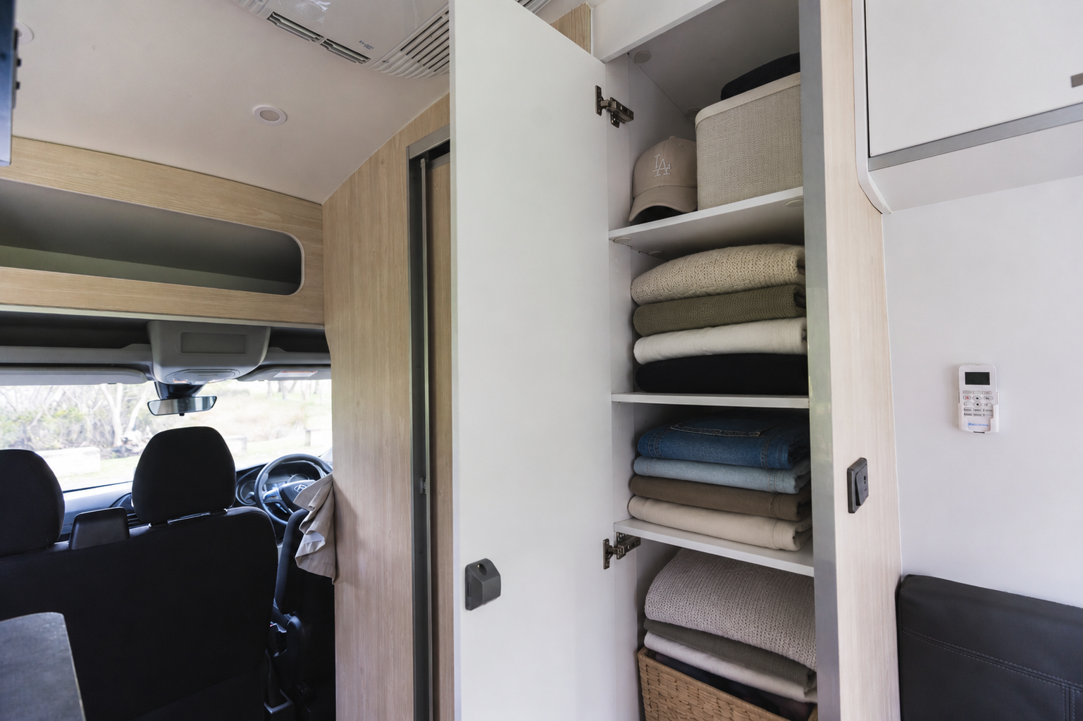 Van interior with open closet showing neatly folded blankets and storage shelves beside driver’s cabin and seating area.