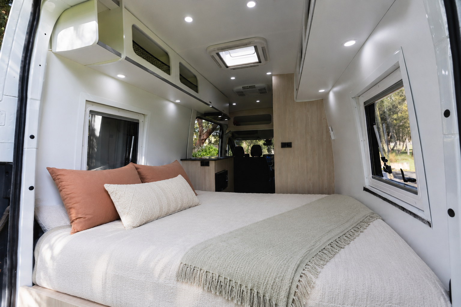 Van interior with large rear bed, overhead storage, side windows, ceiling lights, wooden panel, and kitchen area.