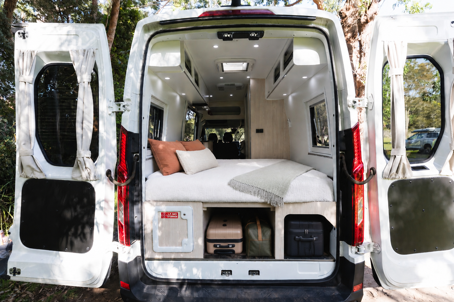 Rear view of van interior with bed, storage compartments, overhead cabinets, and side windows visible.
