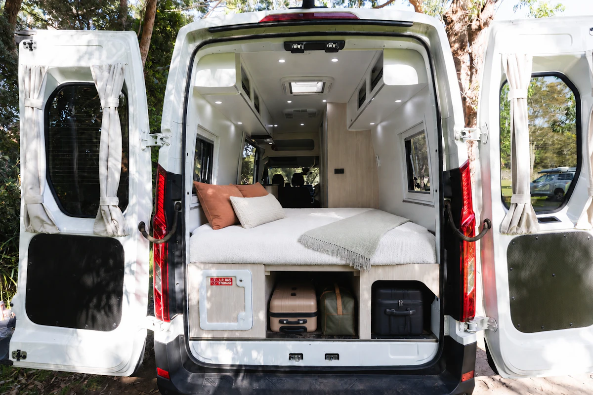 Rear view of van interior with bed, storage compartments, overhead cabinets, and side windows visible.