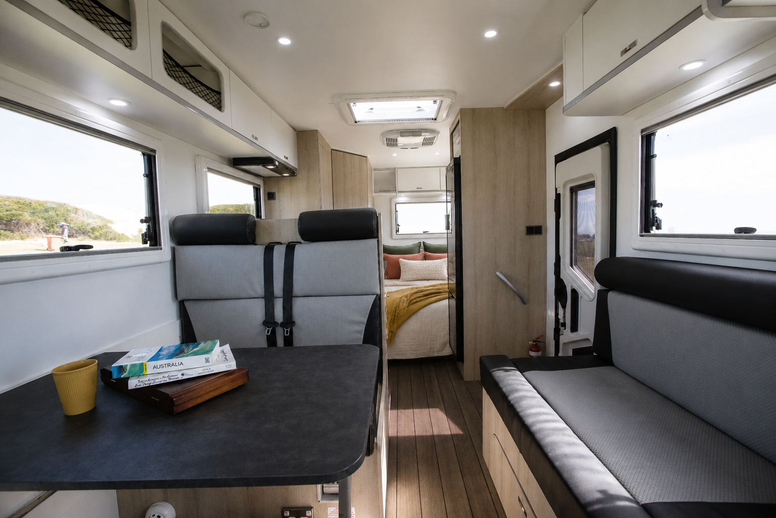 Van interior with dining table, bench seats, overhead storage, windows, and visible rear sleeping area with bed.