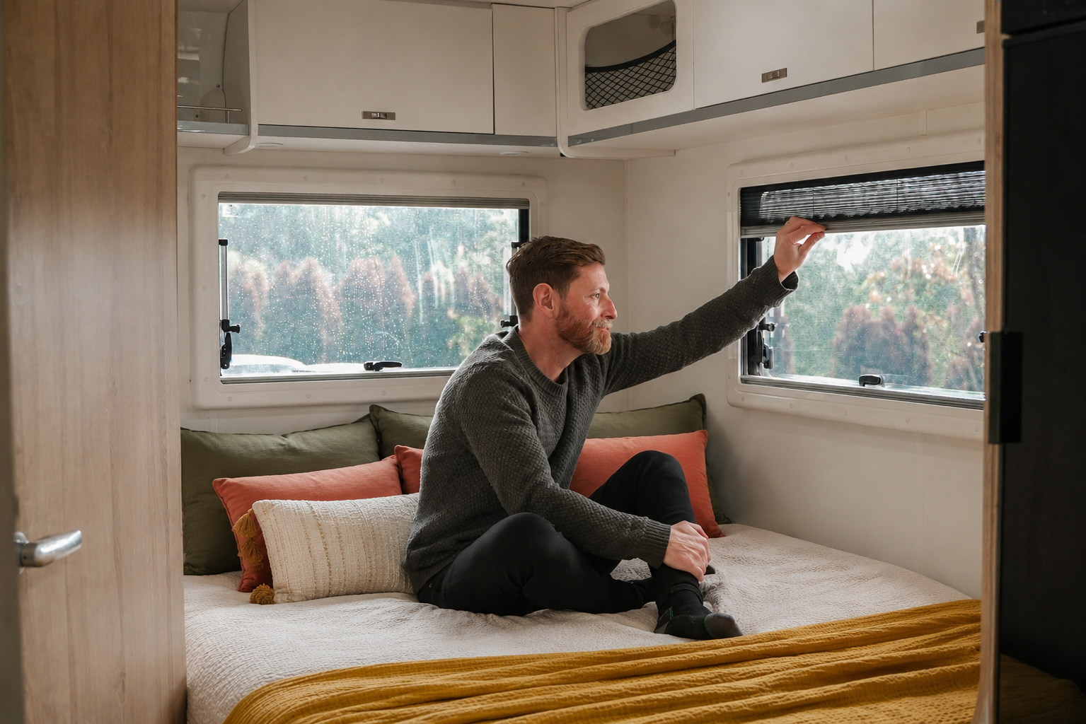 Campervan interior with bed, pillows, windows, overhead cabinets, and a person sitting on the bed.
