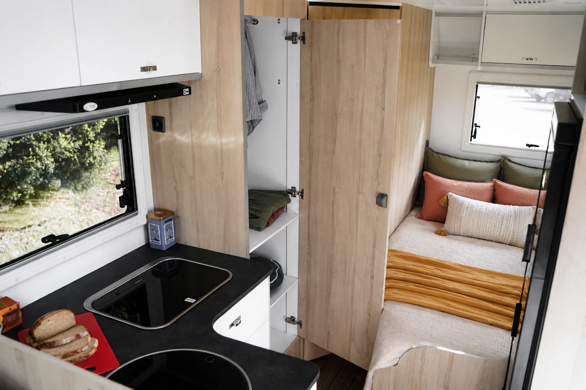 Compact van interior with kitchen, sink, storage cabinets, wardrobe, and rear bed with pillows and blanket.