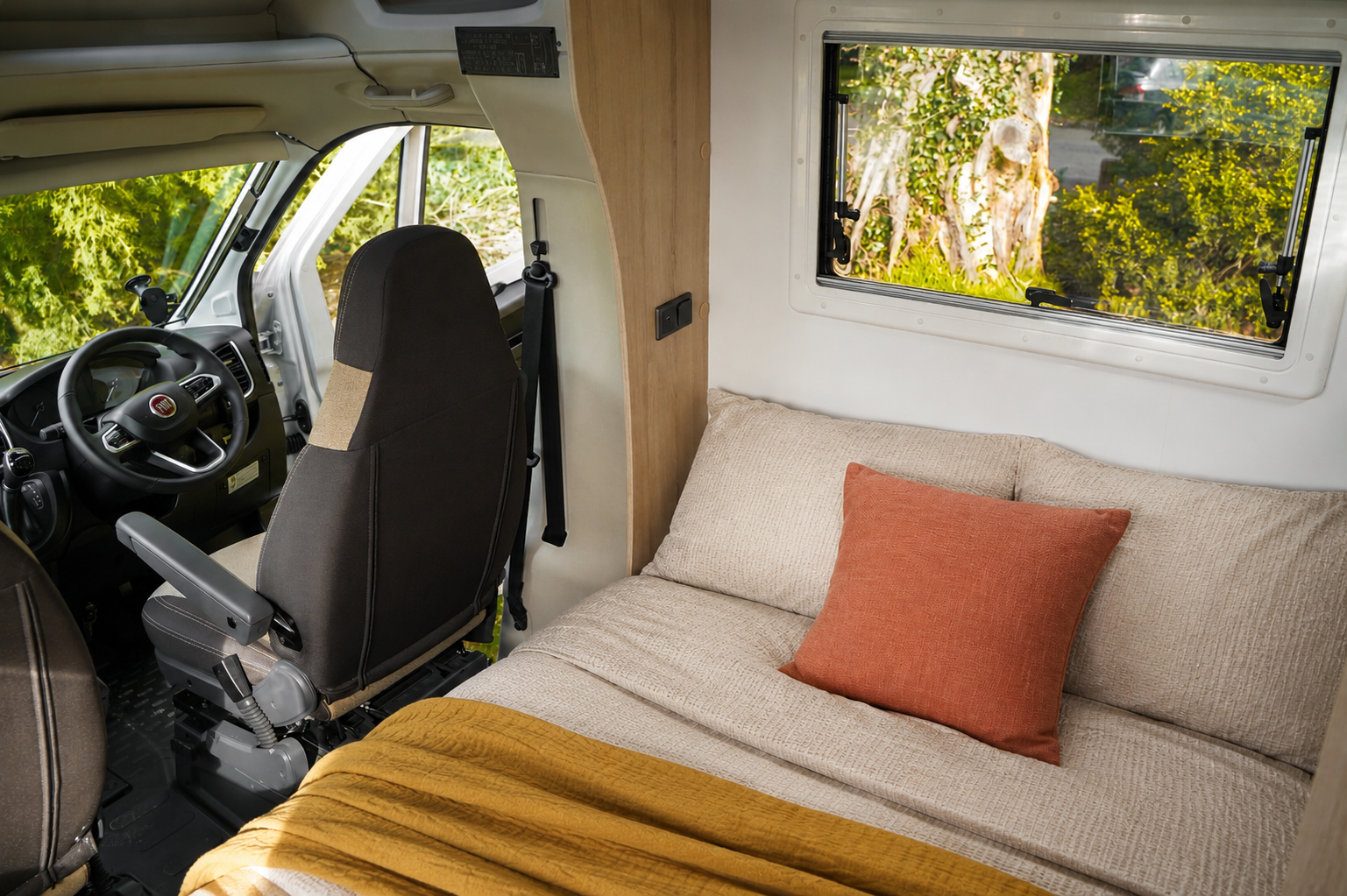 Van interior with front seats, bed, pillow, blanket, side window, and light wood paneling visible.