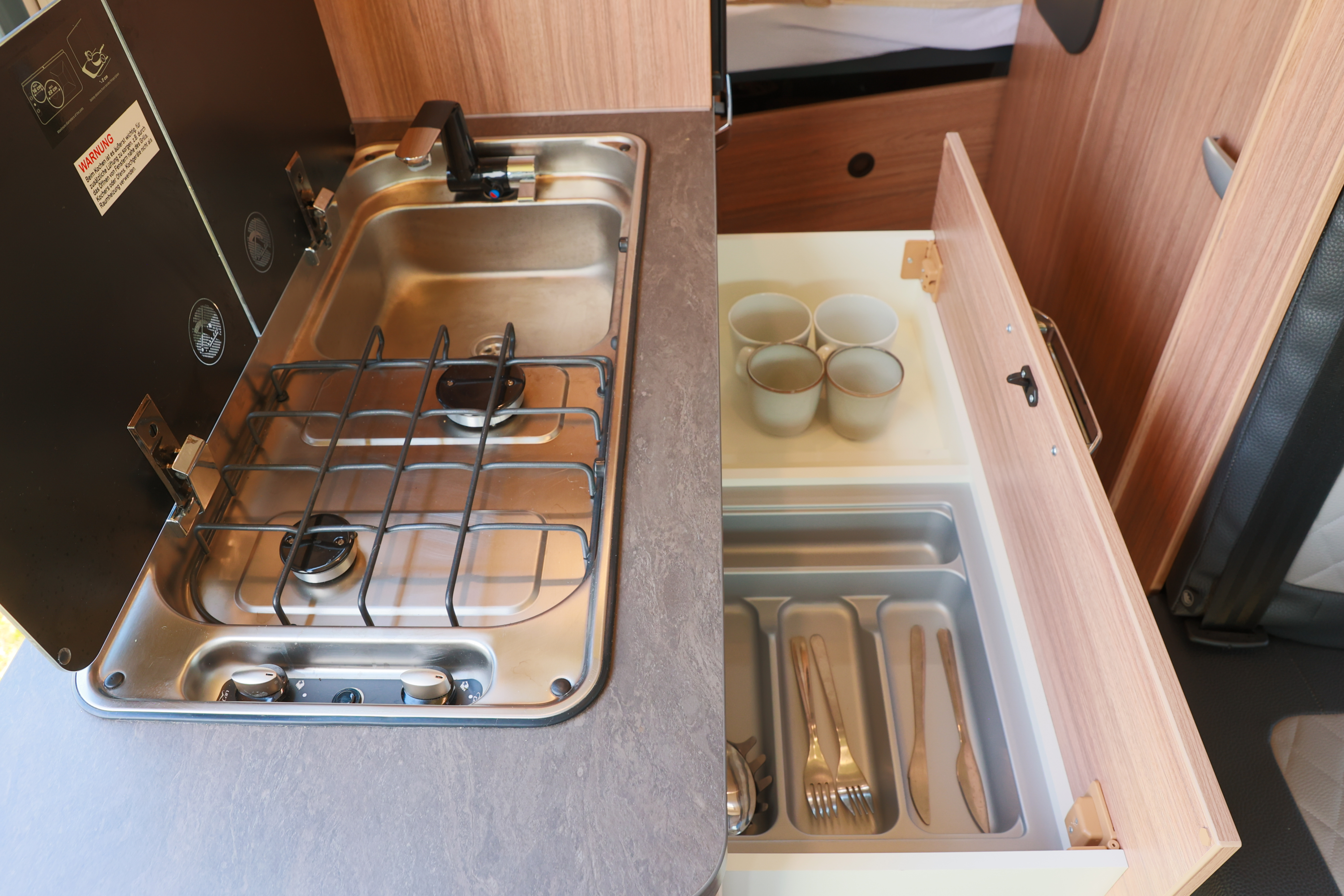 Camper van interior kitchen with two-burner stove, sink, open drawer showing cups and cutlery organizer.