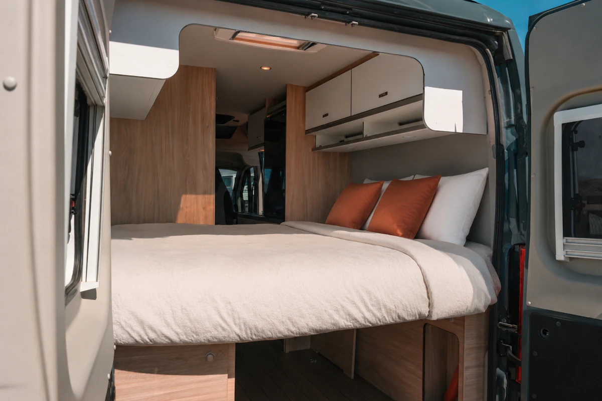 Rear view of van interior showing double bed, overhead storage cabinets, wood paneling, pillows, and ceiling light.