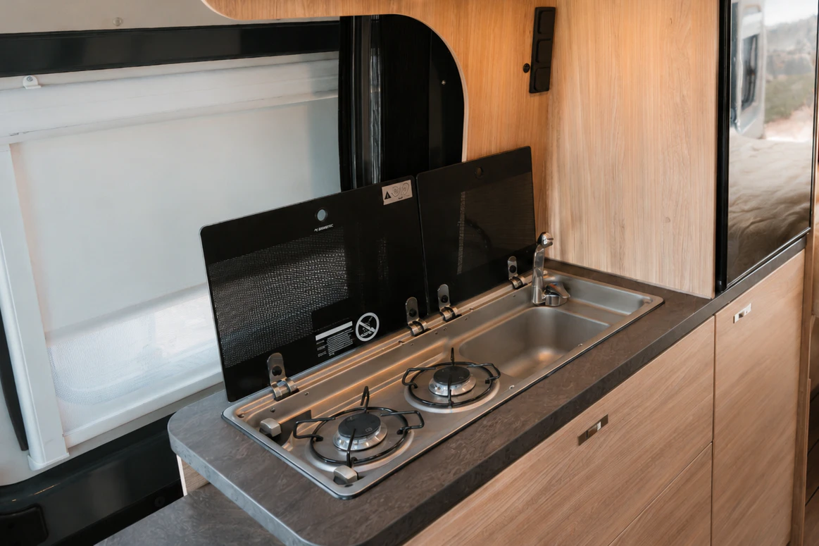 Modern camper van kitchen with two-burner stove, sink, refrigerator, storage cabinets, and visible bed area.