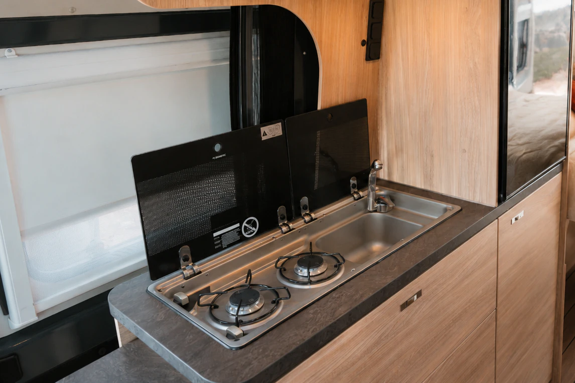 Compact van interior with kitchenette, two-burner stove, sink, refrigerator, upper storage, and visible bed area in background.