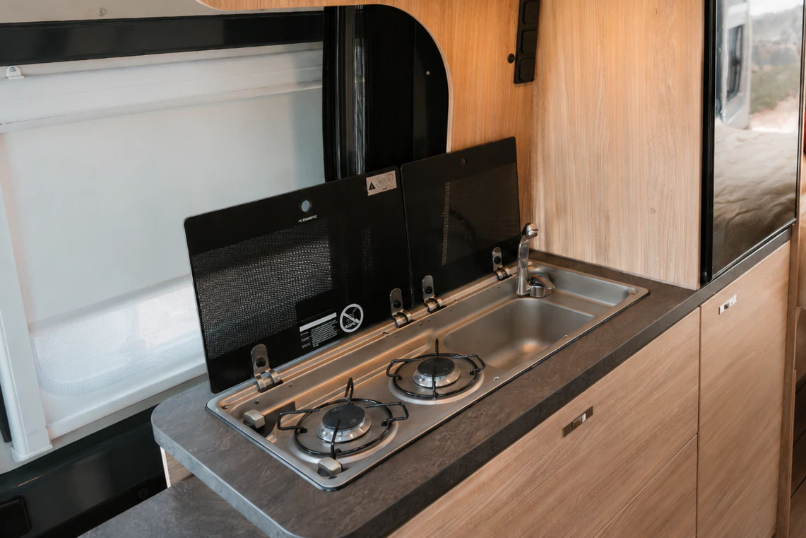 Cocina de furgoneta con dos fuegos, fregadero, nevera, muebles de madera y cama visible al fondo.