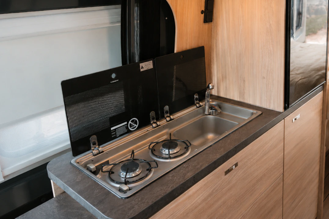 Cozinha de autocaravana com fogão de dois bicos, lavatório, frigorífico, armários e cama ao fundo.
