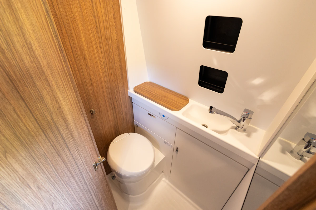 Compact van bathroom with toilet, sink, wooden surfaces, storage cabinets, and wall shelves visible.