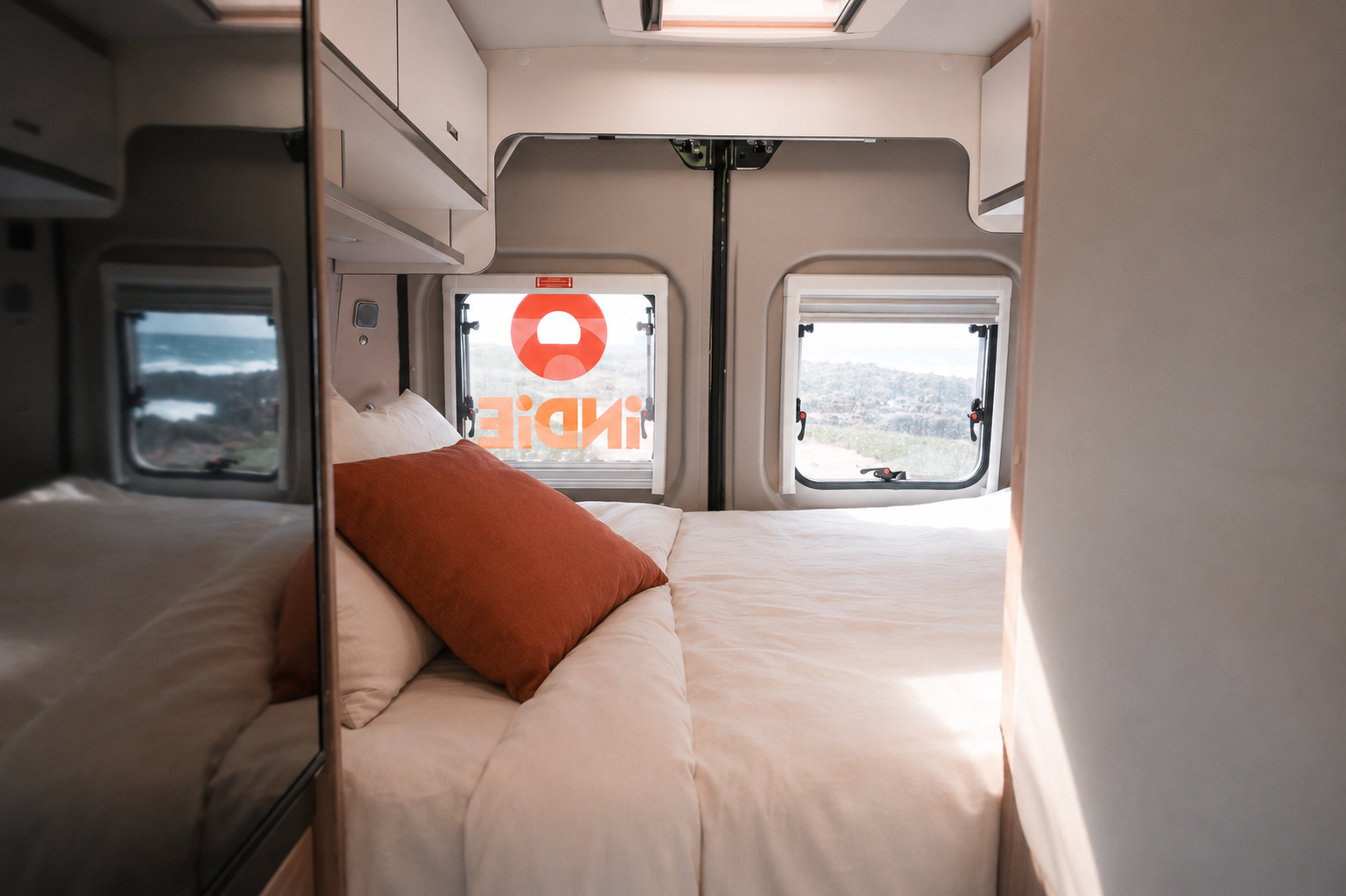 Van interior with a double bed, mirrored cabinet, overhead storage, two rear windows, and pillows.
