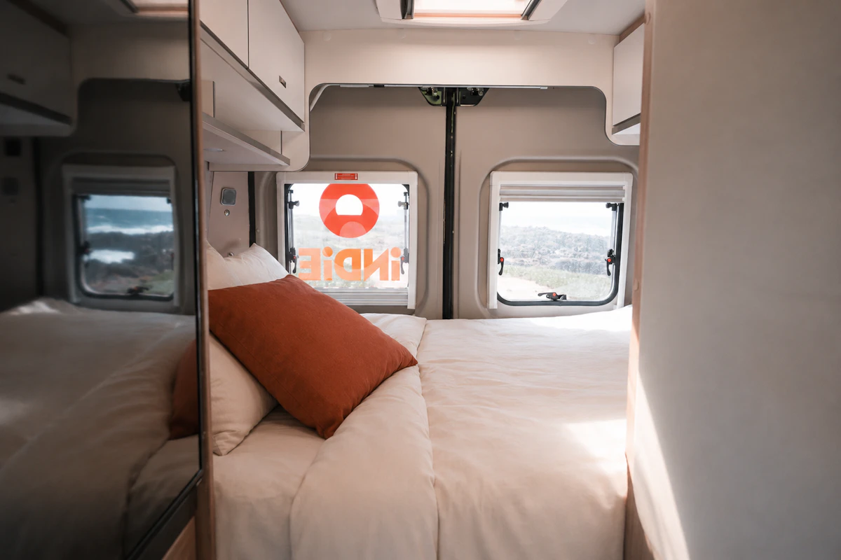 Van interior with a double bed, mirrored cabinet, overhead storage, two rear windows, and pillows.