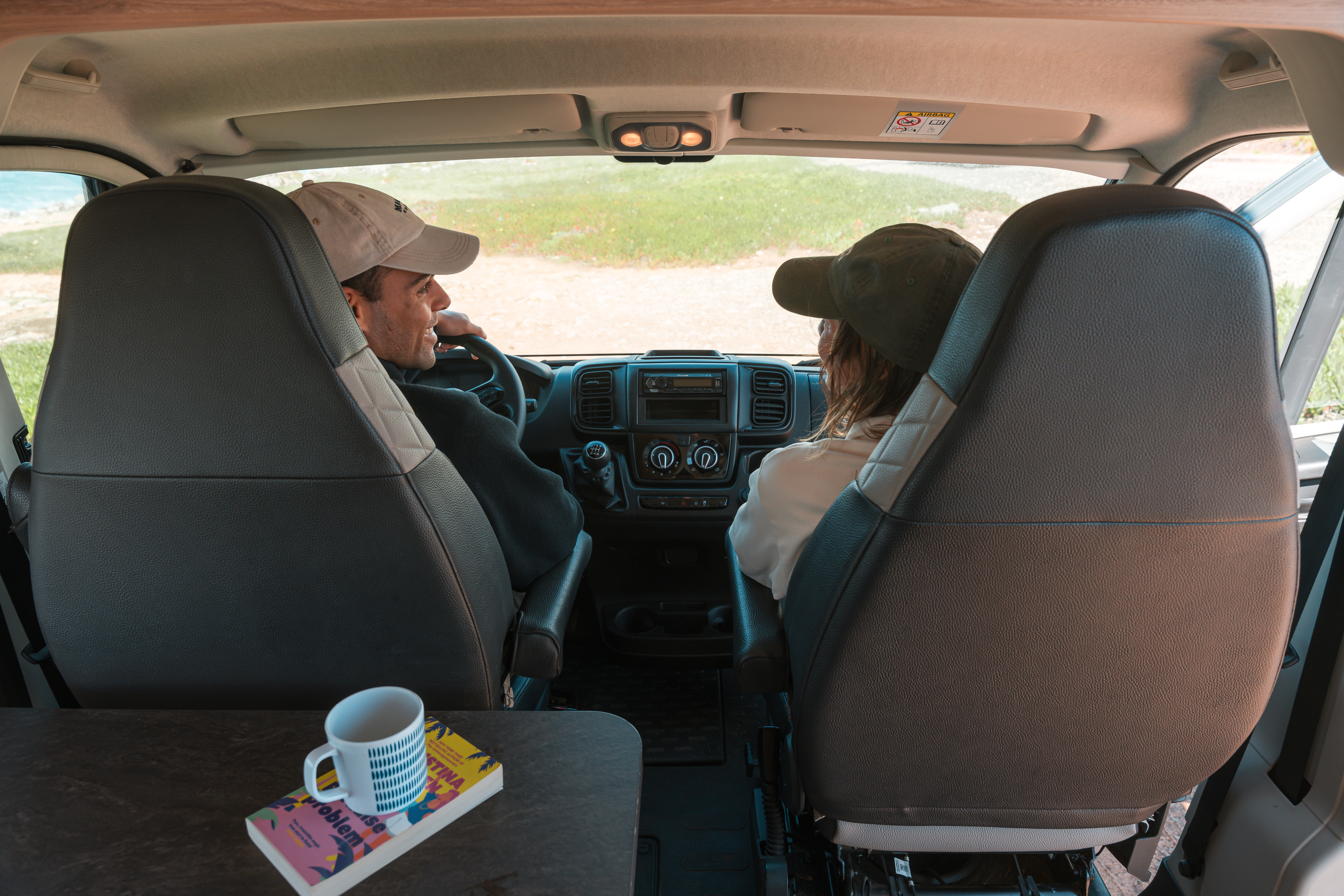 Front interior view of van showing driver and passenger seats, dashboard, table with mug and book.