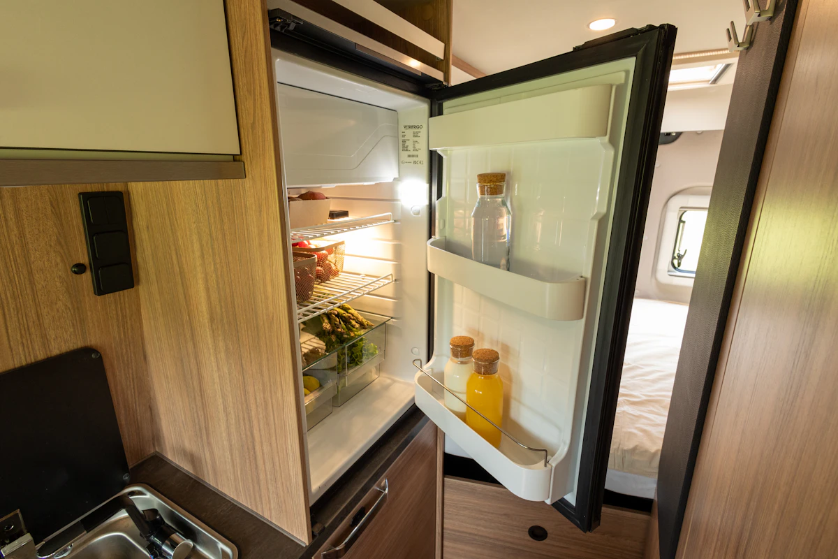 Van interior with open fridge, visible sink, wooden cabinets, and part of a bed in the background.