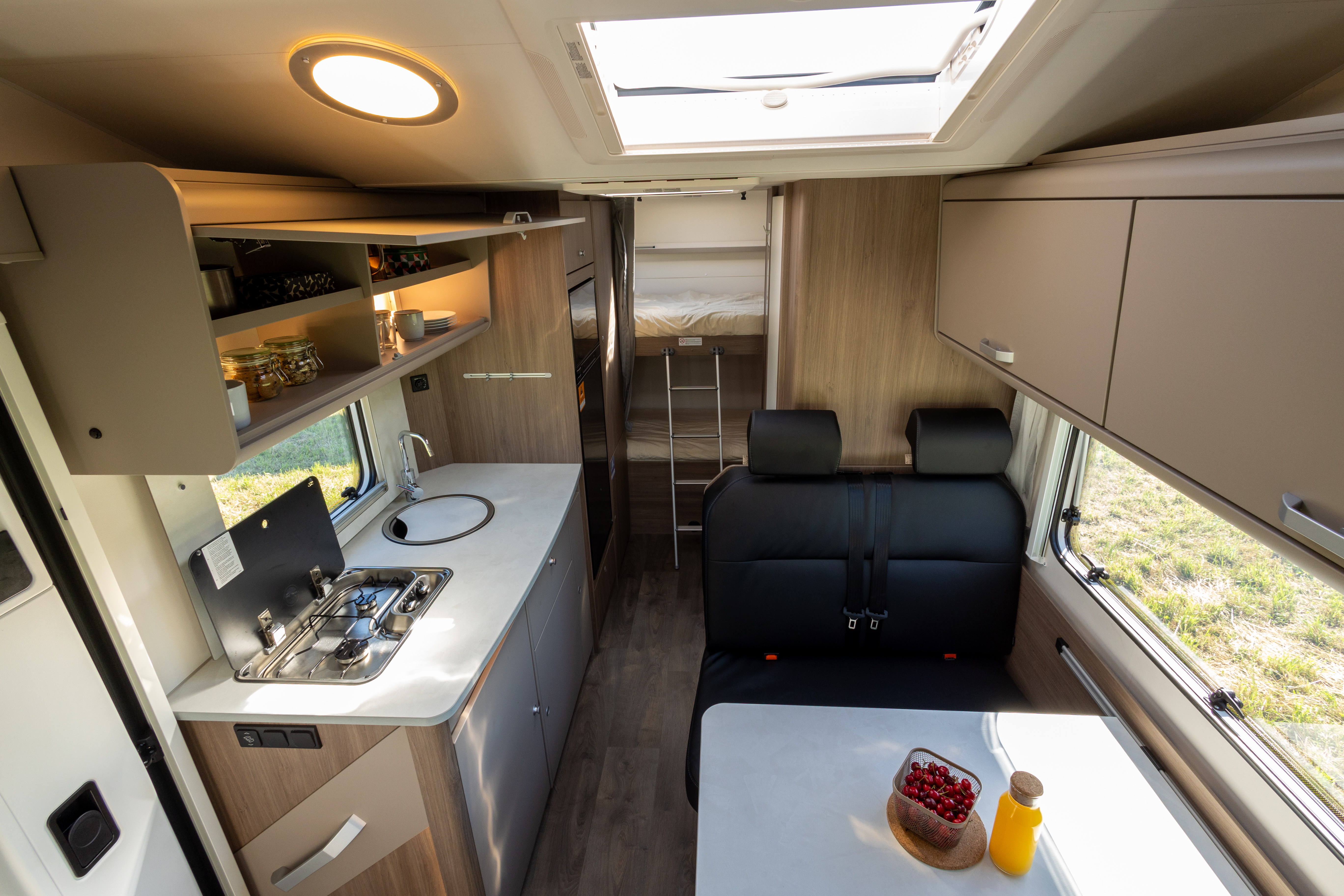 Compact van interior with kitchenette, dining area, overhead cabinets, sofa seating, table, and visible rear sleeping area.