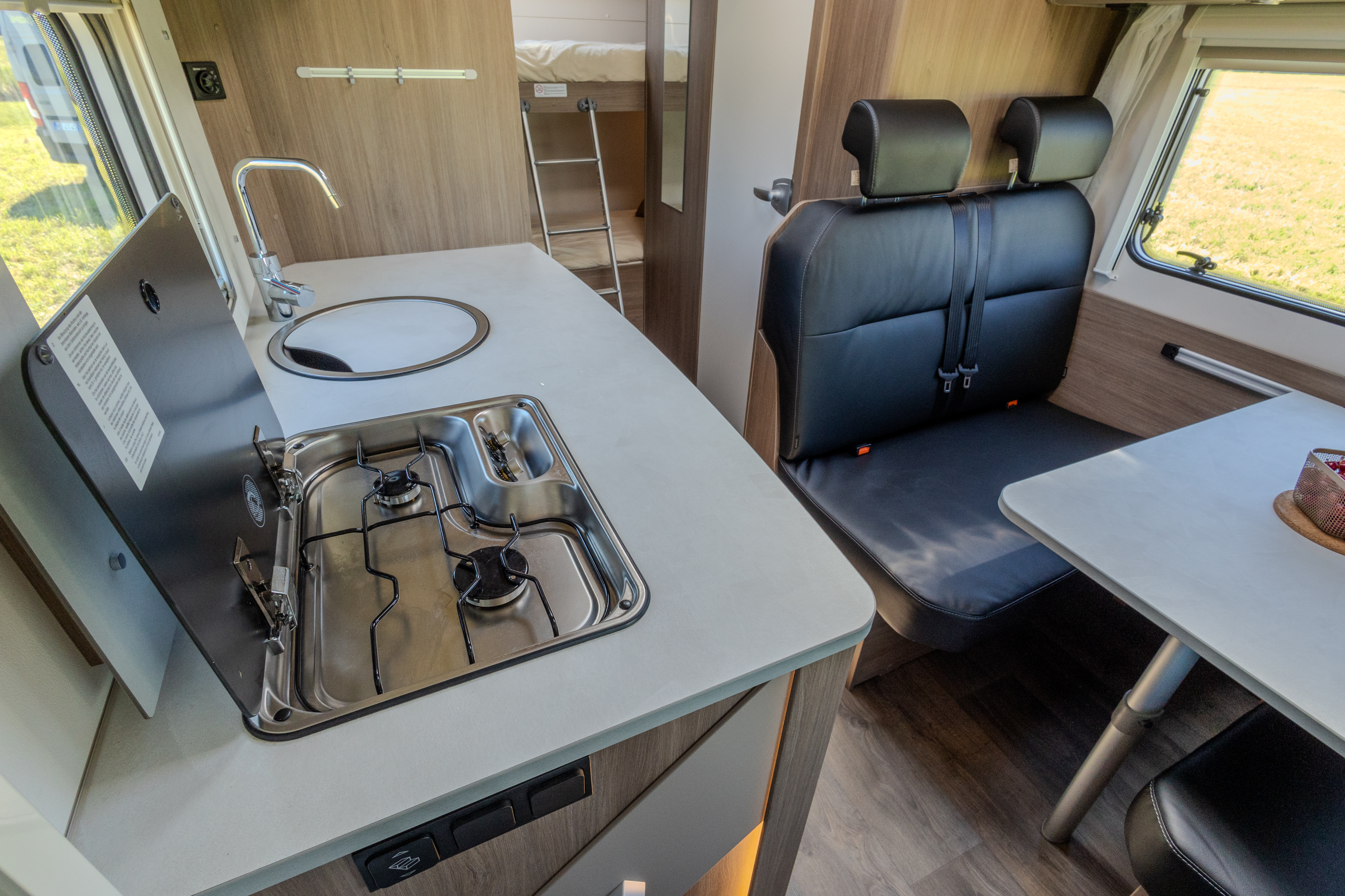 Kitchen with stove and sink, dining table, two-seat bench, window, and visible rear bunk bed inside van.