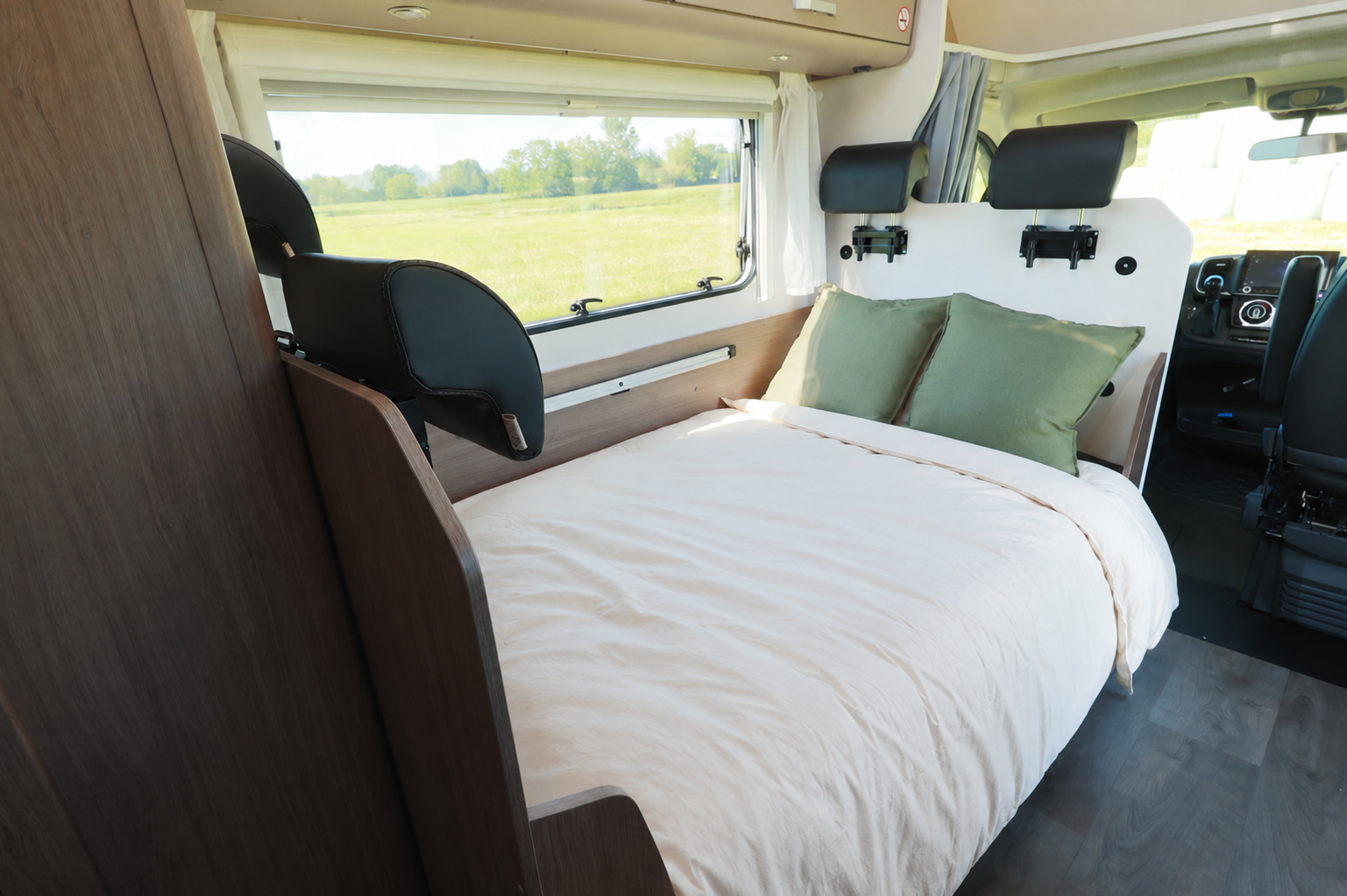 Van interior with a double bed, two pillows, side window, front seats, and dark wood furniture visible.