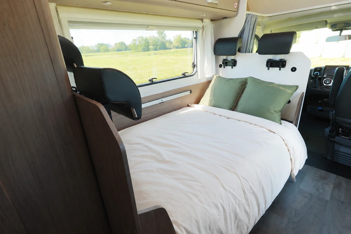 Van interior with a double bed, two pillows, side window, front seats, and dark wood furniture visible.