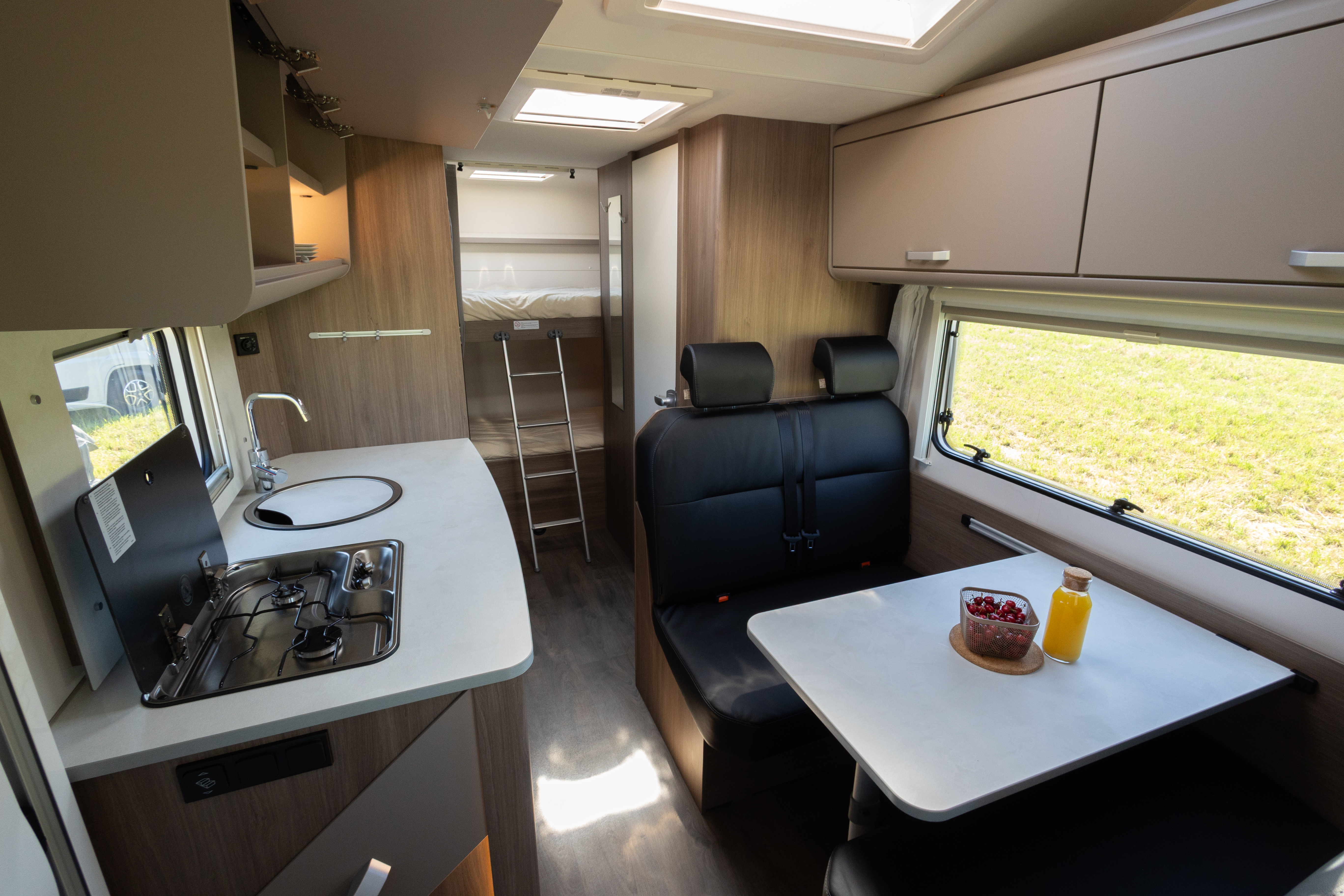 Van interior with kitchen, stove, sink, dining table, bench seating, overhead cabinets, and bed area with ladder.