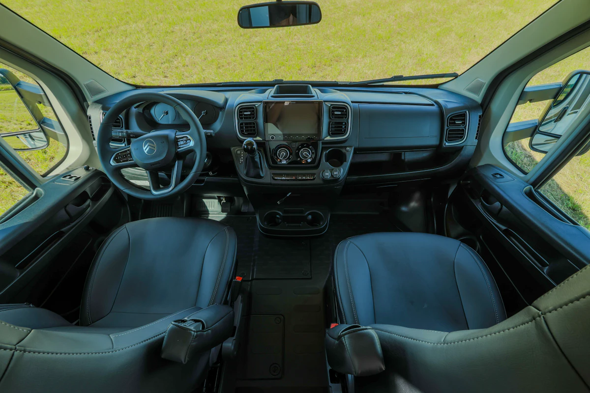 Front interior view of van cockpit with two seats, central touchscreen, dashboard controls, and steering wheel visible.