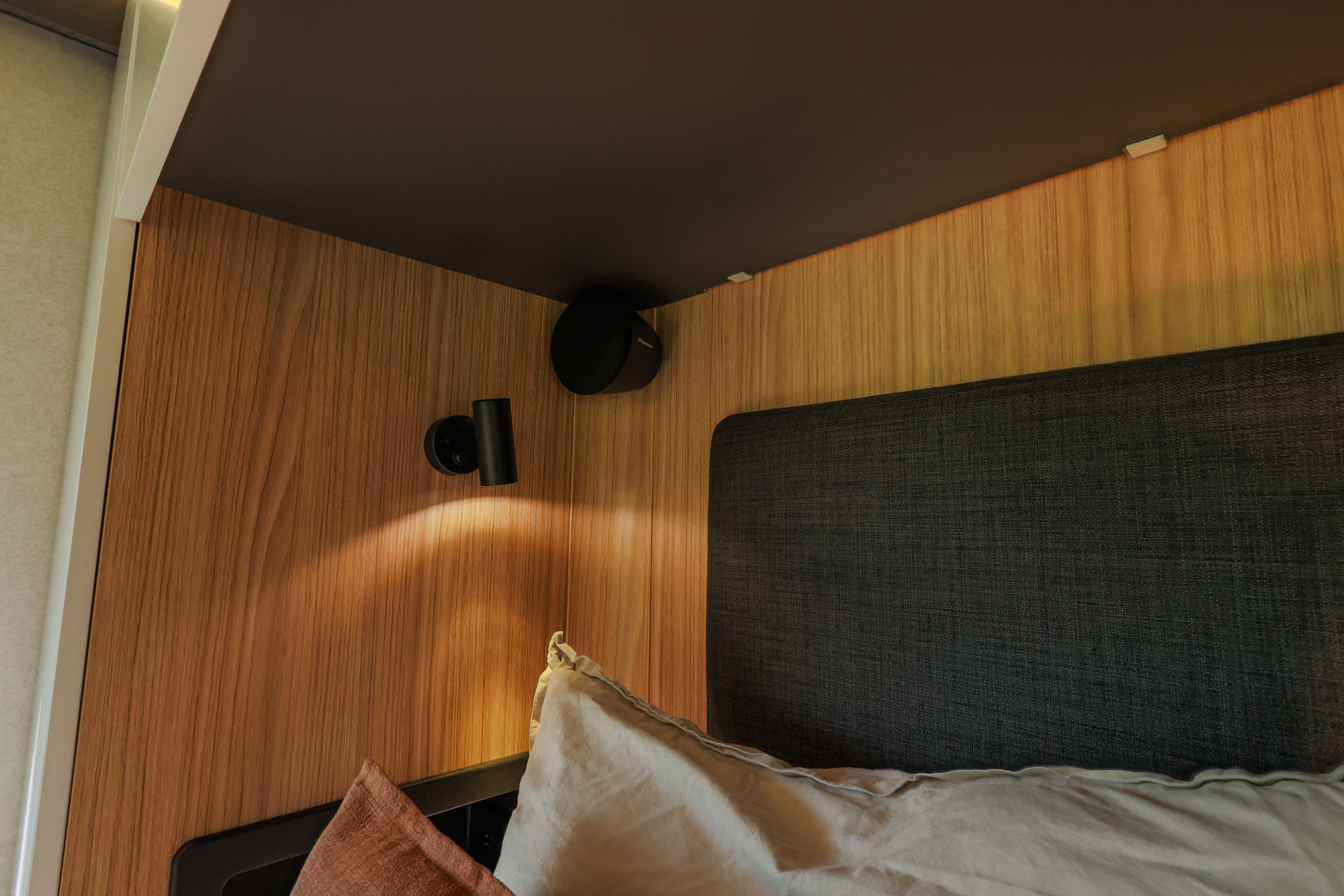 Bed corner in van interior with pillows, wooden wall, reading light, and storage shelf above.