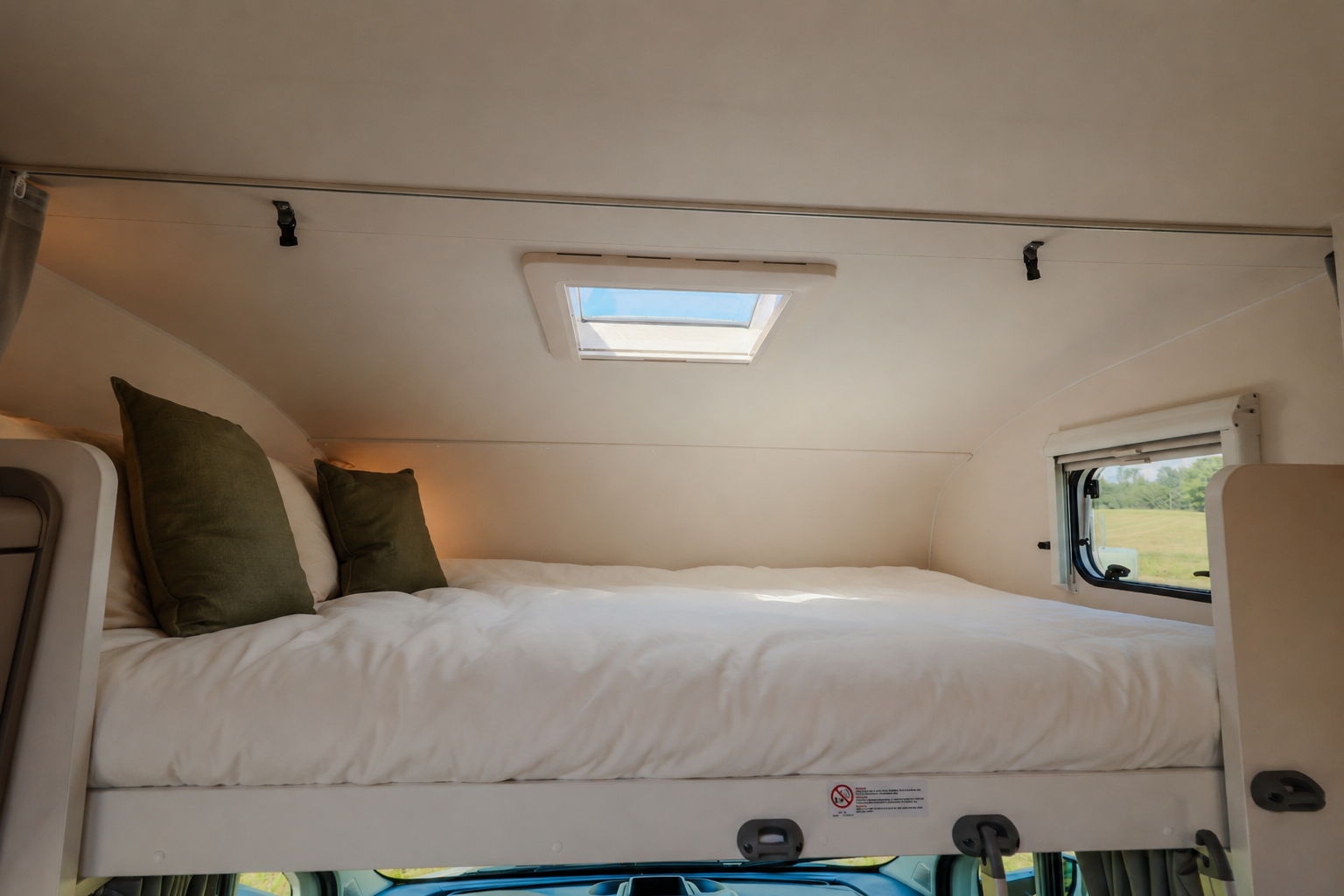 Raised double bed with pillows, roof window above, side window, and storage shelf in the camper van interior.