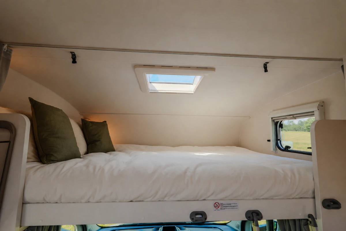 Raised double bed with pillows, roof window above, side window, and storage shelf in the camper van interior.