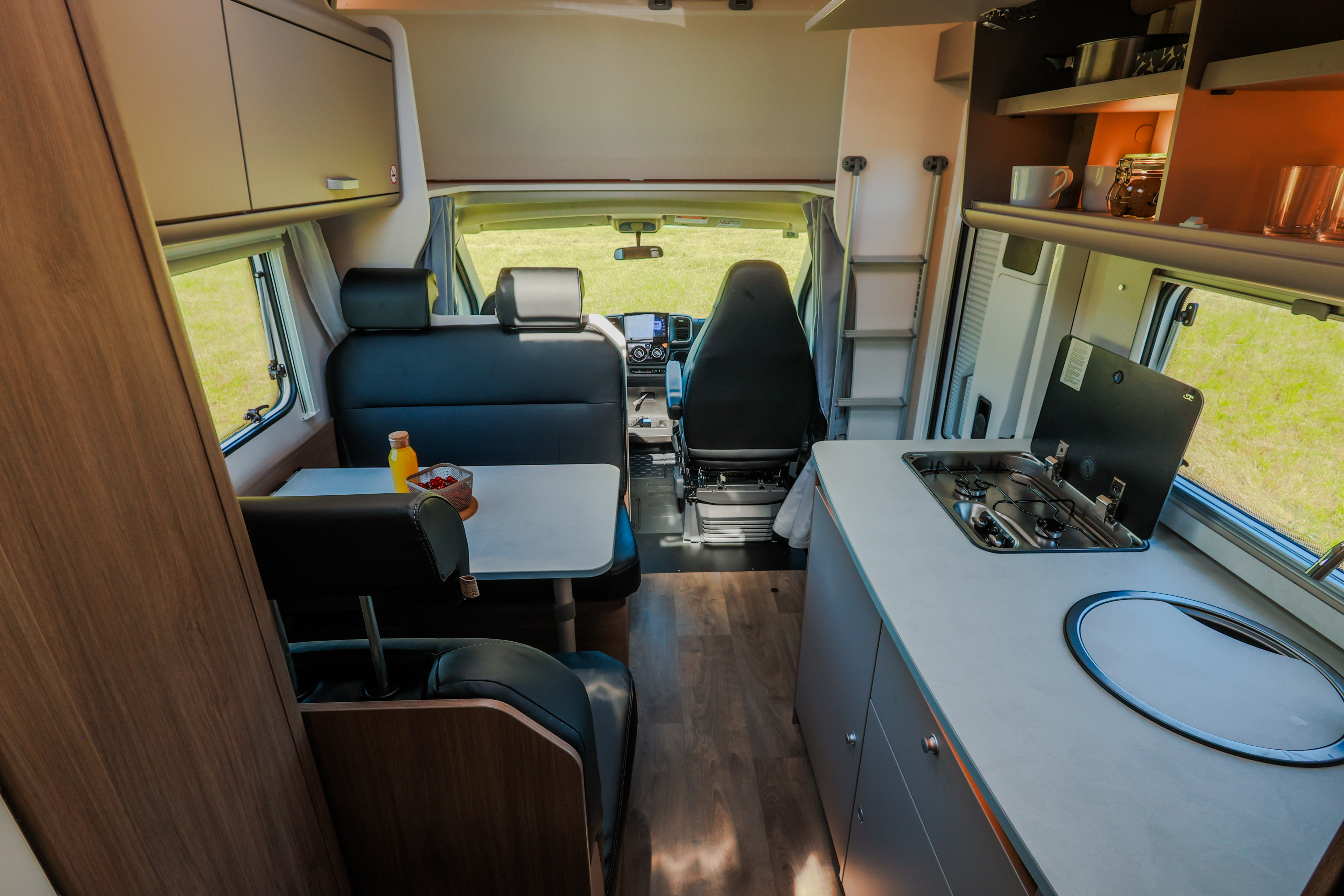 Van interior with kitchen, dining table, bench seats, upper cabinets, and front driving area visible.