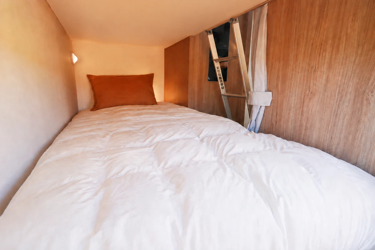 Van interior with single bed, brown cushion, wooden walls, small window, curtain, ladder, and reading light visible.